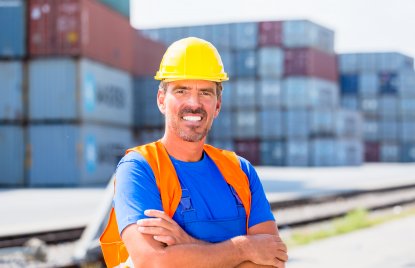 Ein lächelnder Hafenarbeiter mit blauem T-Shirt, orangefarbener Sicherheitsweste und gelbem Sicherheitshelm steht mit verschänkten Armen vor ein Paar Schienen. Im Hintergrund stehen mehrere Wände voller Container.