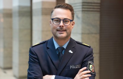 Ein Mann mit kurzen Haaren und Brille in Polizeiuniform und verschränkten Armen schaut lächeld in die Kamera