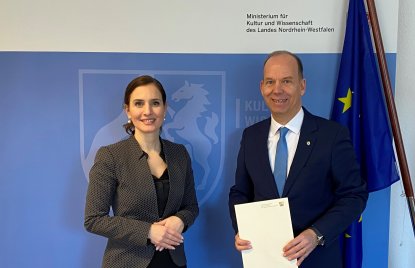 eine Frau und ein Mann stehen vor einem blauen Hintergrund, der Mann hält ein Dokument in beiden Händen