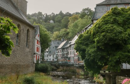 Historische Altstadt mit steinerner Kirche, Fachwerkhäusern und einem Bachlauf, eingerahmt von dichtem grünen Laub. Die idyllische Szene zeigt eine ruhige Flusslandschaft mit Brücke und blühenden Pflanzen.