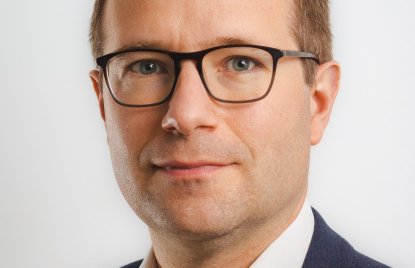 Ein Mann mit kurzen Haaren, Brille, weißen Hemd und blauem Jackett schaut in die Kamera