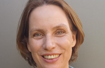 Porträt einer lächelnden Frau mit kurzem, braunem Haar und freundlichem Blick. Sie trägt einen hellblauen Blazer über einem weißen Oberteil und blickt direkt in die Kamera. Der neutrale graue Hintergrund unterstreicht die professionelle und sympathische Ausstrahlung.