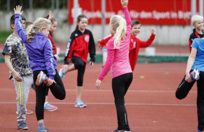 Mehrer Schulkinder machen Dehnübungen auf einem Tartan-Platz.