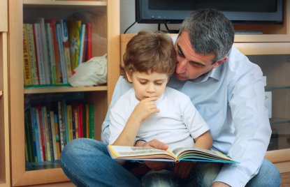 Ein Kind sitzt auf dem Schoß eines Mannes und sie lesen gemeinsam in einem Buch, im Hintergrund ist ein Bücherregal