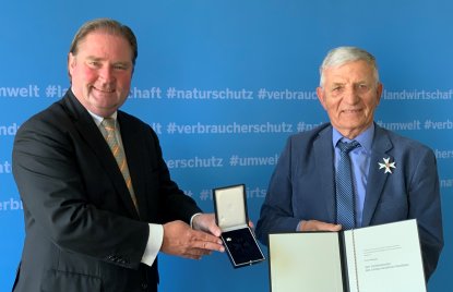 Zwei Männer in Anzügen stehen vor einer blauen Wand und halten einen Orden und ein Dokument