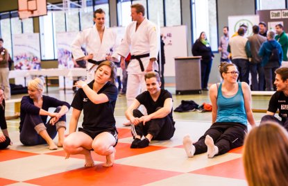 Mehrere Frauen bei Dehnübungen für einen Selbstverteidungskurs in einer Turnhalle. Auf dem Boden grüne und rote Matten im Schachbrettmuster. Im Hintergrund zwei Kampfsportler in weißen Anzügen mit schwarzen Gürteln.