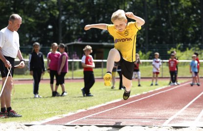 Eine Weitsprunganlage mit roter Tartanbahn. Ein Junge im gelben T-Shirt und schwarzer Hose mitten im Weitsprung. Links daneben steht ein Sportlehrer mit Maßband.