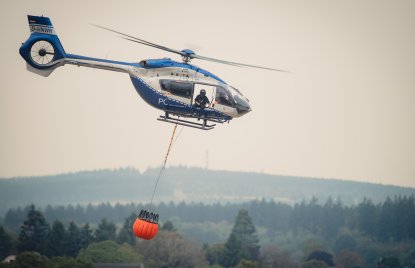 Zu sehen ist ein Löschhubschrauber der Polizei vor einem verrauchten Waldgebiet