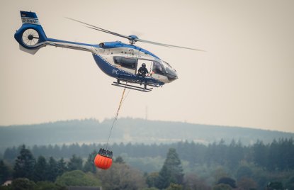 Zu sehen ist ein Löschhubschrauber der Polizei vor einem verrauchten Waldgebiet
