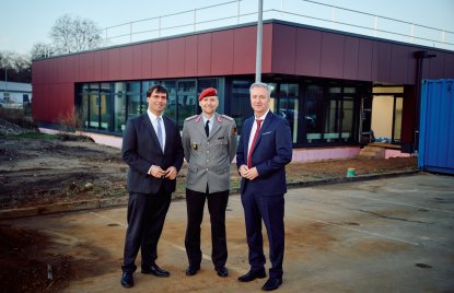 zwei Männer mit Anzug und ein Mann in Bundeswehr-Uniform stehen vor einem roten Gebäude