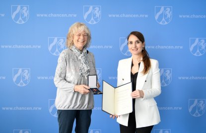 Zwei Frauen stehen vor einer blauen Wand und halten das Bundesverdienstkreuz und eine Urkunde 