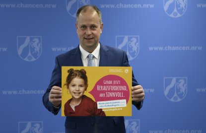 Ein Mann im Anzug steht vor einer blauen Wand und hält ein gelbes Plakat hoch, auf dem ein Kind zu sehen ist