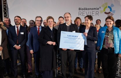 Vor einer Leinwand mit dem Bioökonomie Revier Logo stehen viele Menschen. Die vorderen beiden halten ein Schild hoch.