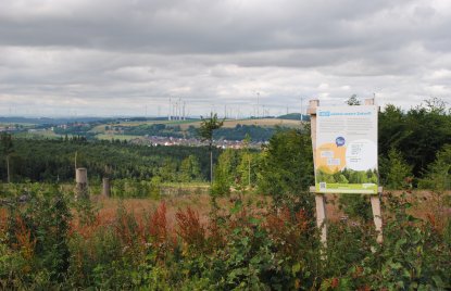 Eine Hinweistafel steht auf einem Hügel an einem Waldrand mit Ausblick in die Ferne