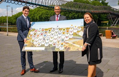 Zwei Männer und eine Frau stehen an einer Uferpromenade und halten ein Schild mit Städte-Wappen hoch