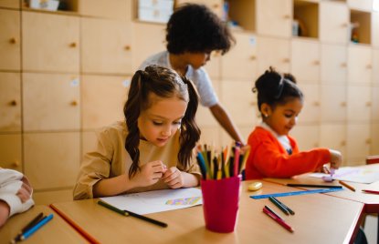 Drei Kinder sitzen an einem Holztisch und beschäftigen sich mit Zeichnen und Malen; im Vordergrund zeichnet ein Mädchen mit Zöpfen, während zwei weitere Kinder im Hintergrund kreativ arbeiten. Auf dem Tisch stehen ein roter Becher mit Buntstiften sowie verschiedene Stifte, Lineale und Zeichenmaterialien.