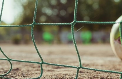 Nahaufnahme eines grünen Fußballnetzes, das im Vordergrund scharf zu sehen ist. Im unscharfen Hintergrund erkennt man einen Ball am rechten Bildrand und eine Spielfläche im Freien.