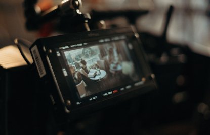 Eine professionelle Filmkamera zeigt auf ihrem Monitor eine Filmszene, in der eine Frau an einem Tisch sitzt und in einem Buch liest. Die warme, stimmungsvolle Beleuchtung und der unscharfe Hintergrund vermitteln einen cineastischen Blick hinter die Kulissen einer Filmproduktion.