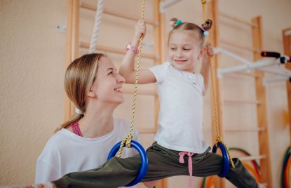 Ein Kind mit weißem T-Shirt und dunkler langer Hose trainiert an Gymnastikringen, während eine Lehrerin es aufmerksam begleitet und motivierend anlächelt.