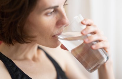 Eine Frau mit schulterlangen, dunklen Haaren und einem schwarzen Tanktop trinkt ein Glas Wasser