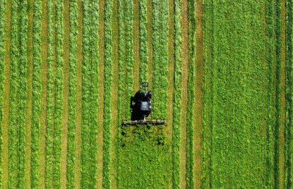 Luftaufnahme eines Traktors, der in parallelen Bahnen ein großes grünes Feld mäht. Die gleichmäßigen Streifen im Gras erzeugen ein grafisches Muster aus verschiedenen Grüntönen.