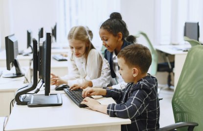 Zwei Mädchen und ein Junge im Grundschulalter sitzen in einem Computerraum gemeinsam vor einem Bildschirm, der Junge tippt etwas auf der Tastatur