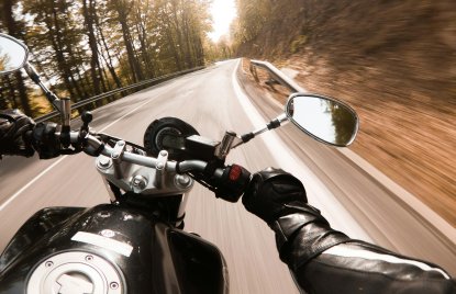 Cockpit eines schwarzen Motorrads während der Fahrt, vom Fahrer sieht man die schwarzen Handschuhe und eine schwarze Jacke. Das Motorrad fährt auf einer Landstraße im Wald bei Sonnenschein.