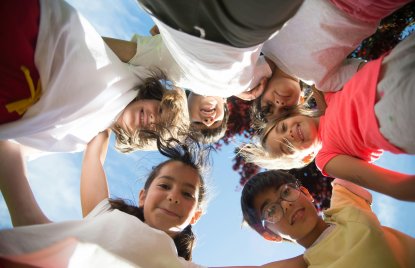 Eine Gruppe von Kindern steht im Kreis und blickt lächelnd nach unten in die Kamera. Die Aufnahme ist von unten gemacht, sodass der Himmel und etwas Baumlaub im Hintergrund zu sehen sind. Die Kinder wirken fröhlich und halten vermutlich zusammen oder lehnen sich zueinander.