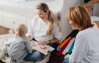 Eine Frau, wie eine weitere Frau mit einem kleinen Kind gemeinsam ein Bilderbuch anschaut. Die drei sitzen in einer hellen, wohnlichen Umgebung und wirken aufmerksam miteinander im Gespräch.
