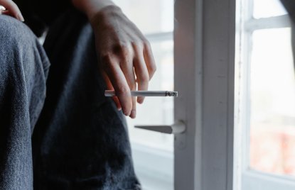 Eine Person sitzt neben einer Glastür und hält eine brennende Zigarette zwischen den Fingern. Die Hand ist im Vordergrund zu sehen, im Hintergrund sind Jeansstoff und ein Türgriff sichtbar. Tageslicht fällt durch das Fenster.