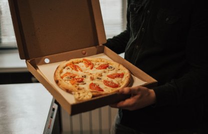 Ein Mensch mit schwarzer Kleidung (Kopf ist nicht sichtbar) hält eine Packung eines Pizza-Lieferdienstes in der Hand, die offen ist und eine Pizza mit Tomaten zeigt. Hintergrund ist eine Heizung und ein kleiner Kühlschrank erkennbar.