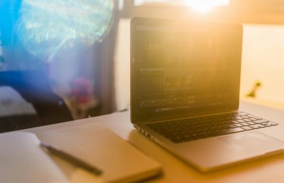 auf einem hellen Schreibtisch liegen ein Notizbuch mit einem Stift links und recht ein aufgeklappter Laptop, auf dem verschwommen ein dunkler Bildschirm mit Zahlen zu erkennen ist. Im Hintergrund scheint durch das Fenster die Sonne und eine Spiegelung der Sonne verdeckt den restlichen Hintergrund.