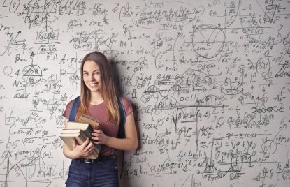 Eine junge Frau steht in Jeans-Latzhose und einem rötlichem T-Shirt mit mehreren Büchern in den Armen vor einer Tafel voller handschriftlicher mathematischer Formeln und Diagramme.