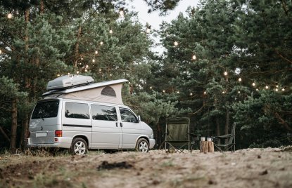 ein silberner VW Bus mit ausgeklapptem Dach und Dachbox steht auf einem Campingplatz im Wald, daneben zwei Klappstühle und zwei Becher, im Hintergrund eine Lichterkette und der Wald