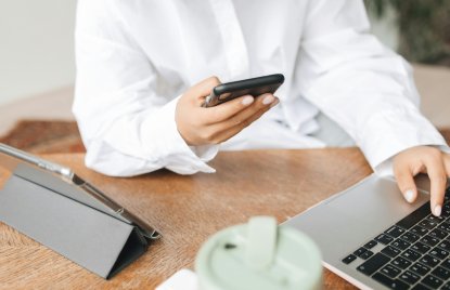 Person in weißer Bluse sitzt am Schreibtisch und nutzt gleichzeitig Smartphone und Laptop. Auf dem Tisch stehen außerdem ein Tablet in Schutzhülle und ein Becher – modernes Arbeiten im Homeoffice oder Büro.
