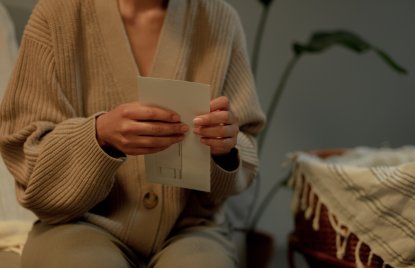 Person in beigem Strickpullover hält einen Brief in den Händen und sitzt auf einem Sofa. Im Hintergrund steht eine Pflanze, rechts ist ein Korbstuhl mit einer Decke zu sehen.