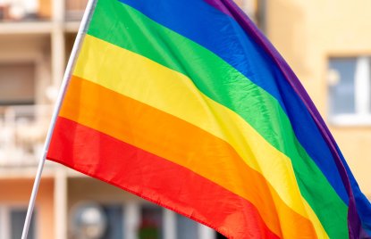 Fahne in Regenbogenfarben, im Hintergrund leicht verschwommen ein gelbes Mehrfamilienhaus