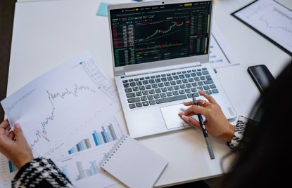 Person analysiert Börsencharts auf einem Laptop und vergleicht diese mit ausgedruckten Diagrammen. Die Szene zeigt die Auswertung von Finanz- und Marktdaten.