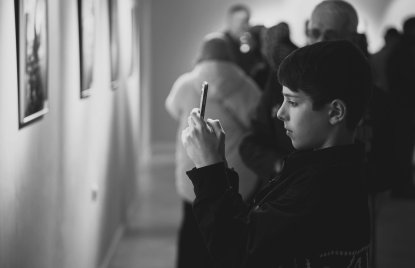 Ein Junge steht in einer Galerie und fotografiert konzentriert ein Kunstwerk mit seinem Smartphone. Im Hintergrund sind weitere, unscharfe Besucher zu sehen, die sich ebenfalls die Ausstellung ansehen. Die Szene ist in Schwarz-Weiß gehalten und wirkt ruhig und aufmerksam.