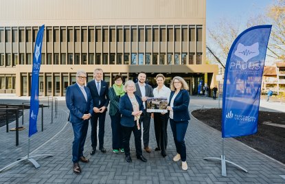 Wolfgang Feldmann, Dr. Dirk Günnewig, Gabriele Willems, Simone Probst, Prof. Dr. Matthias Bauer, Ministerin Ina Brandes und Prof. Dr. Christine Silberhorn halten ein Bild in der Hand vor dem Gebäude der PhoQS. Das Bild, ein Geschenk vom BLB NRW, dient als elektronischer Schlüssel für das Gebäude.