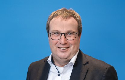 Ein Mann mit braunen kurzen Haaren, Brille, weißem Hemd und schwarzem Jackett lächelt in die Kamera