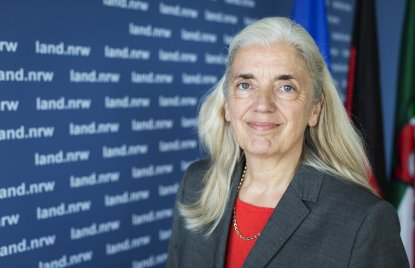 Ministerin Isabel Pfeiffer-Poensgen lächelnd vor EU-, Deutschland- und NRW-FLaggen