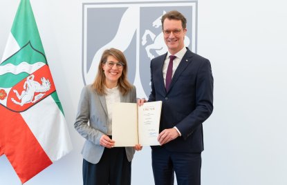 Rechts neben der NRW-Fahne steht Verena Schäffer mit langen blonden Haaren und Brille im grauen Sakko und weißem Hemd lächelnd neben MP Wüst im schwarzen Anzug mit roter Krawatte