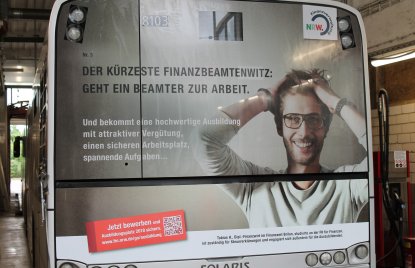 Ein Plakat auf der gesamten Rückseite eines Buses aufgeklebt. Ein Mann mit Brille rauft sich die Haare. Links daneben der Witz: "Geht ein Beamter zur Arbeit..."