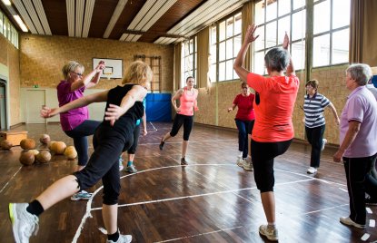Neun Damen in Sportbekleidung stehen in der Mitte einer Sporthallte im Kreis und machen Aufwärmübungen.