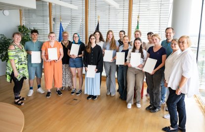 17 Menschen stehen vor drei Flaggen, einige halten eine Urkunde in den Händen