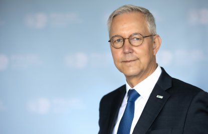 Oberkörper eines Mannes mit grauen Haaren, runder Brille und einem schwarzen Anzug mit dunkelblauer Krawatte und weißem Hemd. Im Hintergrund ist unscharf eine Logowand der Polizeibehörde Dortmund zu erkennen, weiße Schrift auf hellblauem Untergrund.