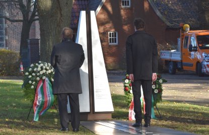 Zwei Männer stehen vor einem Gedenkstein, der mit zwei Blumenkränzen geschmückt ist
