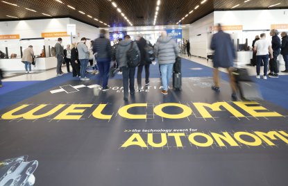 Eingangsbereich der XPONENTIAL, auf dem Boden steht in gelber Schrift "WELCOME" und man sieht einige Menschen, die in die Messehalle gehen. Der Hintergrund ist nur unscharf erkennbar mit weißen Wänden und Messeständen.