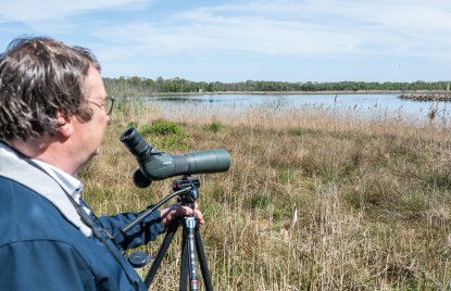 Minister Krischer steht am Rand eines Schilfgebiets und beobachtet mit einem Spektiv auf einem Stativ eine Wasserfläche. Im Hintergrund sind ein See, Vögel und eine flache, bewaldete Landschaft unter blauem Himmel zu sehen.
